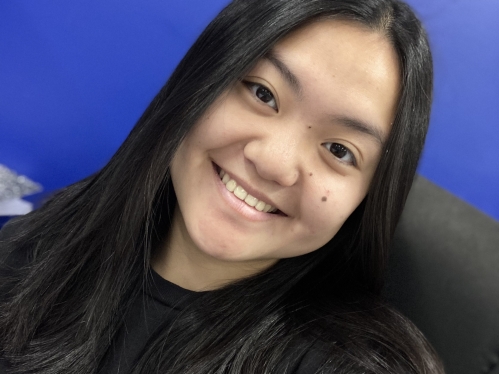 Young woman poses against a blue background. She has black hair and is wearing a black shirt.